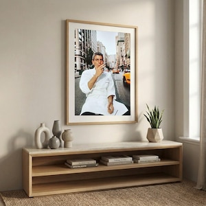 a framed photograph of a man in a white chef's coat, sitting on a bench in a city street, with a cityscape in the background. The photograph is hung on a wall above a wooden entertainment center with shelves below it, which also holds a potted plant and some books.