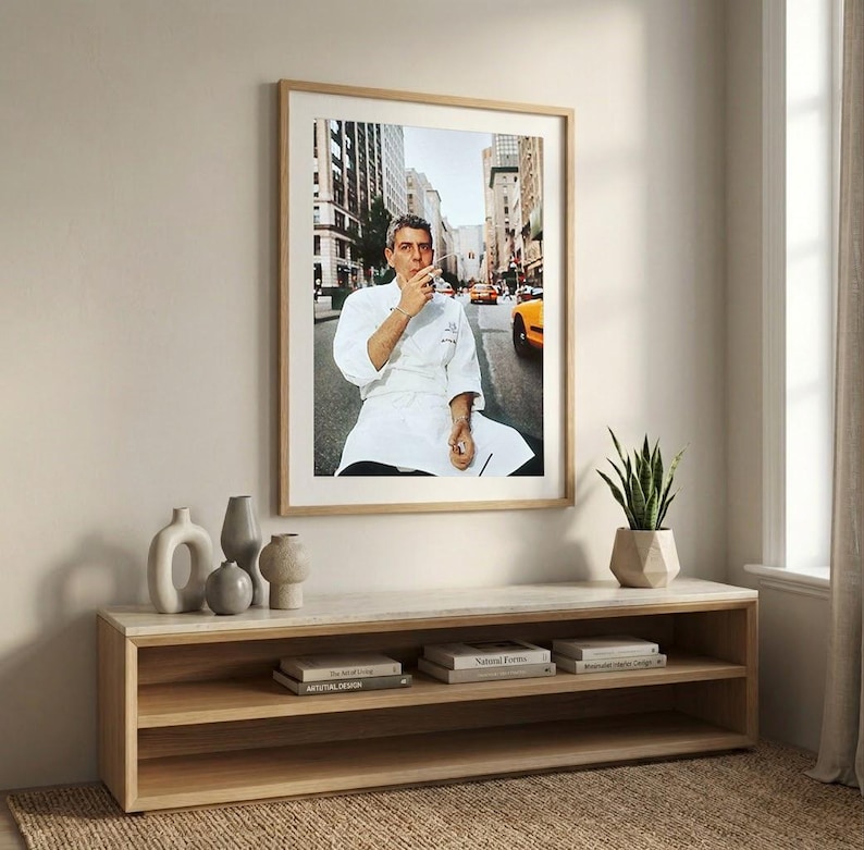 a framed photograph of a man in a white chef's coat, sitting on a bench in a city street, with a cityscape in the background. The photograph is hung on a wall above a wooden entertainment center with shelves below it, which also holds a potted plant and some books.