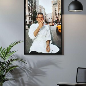 a framed photograph of a chef sitting on a bench in a city street, with a potted plant and a black chair visible in the foreground.