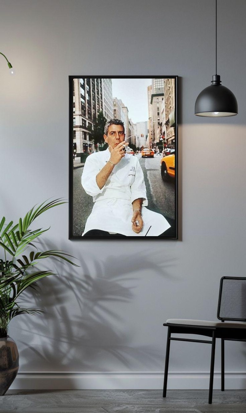 a framed photograph of a chef sitting on a bench in a city street, with a potted plant and a black chair visible in the foreground.