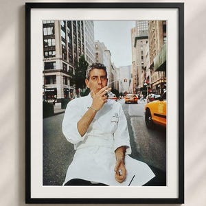 A framed photograph of a chef in a white uniform sitting on a bench in a city street, smoking a cigarette.