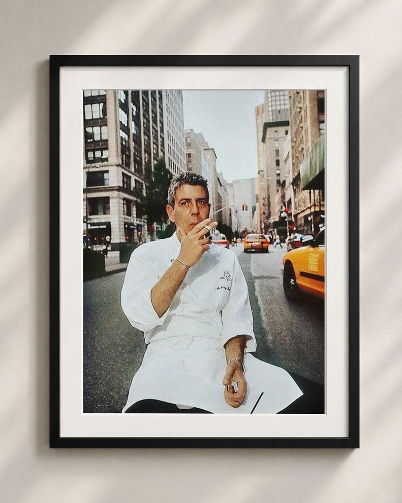 A framed photograph of a chef in a white uniform sitting on a bench in a city street, smoking a cigarette.