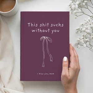 May include: A dark purple hardcover journal featuring the white text "This shit sucks without you" at the top, a simple white line drawing of a ribbon in the center, and the white text "I miss you, mom" printed at the bottom of the front cover.