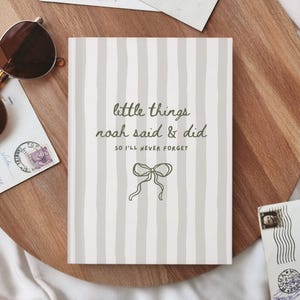 Op de afbeelding: Een rechthoekig notitieboek met een lichtgrijze en witte verticaal gestreepte kaft. De tekst luidt "little things noah said & did SO I'LL NEVER FORGET" in donkergroen, vergezeld van een eenvoudige lijntekening van een strik.