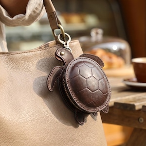 May include: A dark brown leather coin purse shaped like a turtle with a hexagonal shell pattern and a zipper closure. The turtle accessory is attached to a beige leather handbag by a brass-colored metal lobster clasp and a short leather strap.