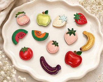 Puede incluir: Una colección de doce pequeños broches de cuero con formas de frutas coloridas. El conjunto incluye representaciones de fresas, manzanas, rodajas de sandía, un plátano, un tomate y una berenjena, cada uno con costuras visibles y hojas verdes.