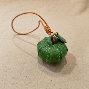 Puede incluir: Un peque&ntilde;o dije de calabaza de cuero verde texturizado con costuras verdes visibles, un tallo rojo oscuro y una peque&ntilde;a hoja verde. El dije est&aacute; unido a un cord&oacute;n de cuero marr&oacute;n claro con un lazo anudado para colgar o sujetar.
