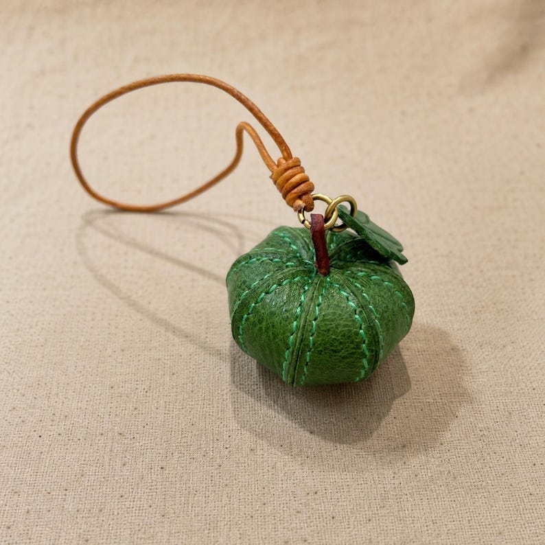 Puede incluir: Un peque&ntilde;o dije de calabaza de cuero verde texturizado con costuras verdes visibles, un tallo rojo oscuro y una peque&ntilde;a hoja verde. El dije est&aacute; unido a un cord&oacute;n de cuero marr&oacute;n claro con un lazo anudado para colgar o sujetar.