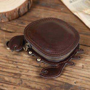 Könnte beinhalten: Eine dunkelbraune Münzbörse aus Leder in Krabbenform mit einem abgerundeten Hauptfach, Reißverschluss, Metallbeschlägen und sichtbaren Nähten. Das Design umfasst hervorstehende Krabbenscheren und Beine an den Seiten.