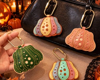 Puede incluir: Cuatro llaveros de cuero con forma de calabaza y mosquetones dorados. Uno es verde oscuro, otro combina tonos pastel amarillo, rosa y azul, uno es rosa sólido y otro presenta un diseño multicolor con segmentos en azul, rosa y amarillo.