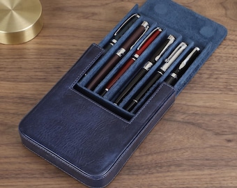 Puede incluir: Un estuche para bolígrafos de cuero azul marino con un forro interior azul suave, diseñado para guardar cinco instrumentos de escritura. El estuche está abierto y muestra cinco bolígrafos distintos con colores de cuerpo variados, incluyendo negro, marrón, rojo y plata.