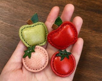 Puede incluir: Cuatro pequeños adornos de cuero hechos a mano con forma de fruta. El conjunto incluye una manzana verde claro, una manzana roja brillante, una fruta redonda de color rosa pálido y un tomate rojo vibrante, cada uno con una hoja de cuero verde y costuras.