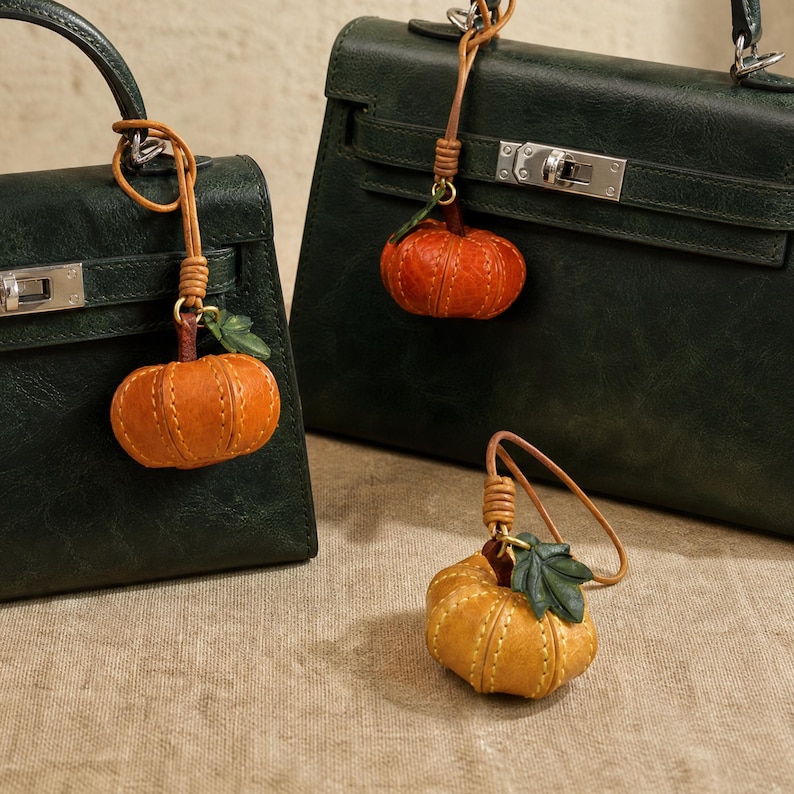 Puede incluir: Dos bolsos de mano de cuero verde oscuro con herrajes plateados, cada uno con un dije de calabaza de cuero naranja y una peque&ntilde;a hoja verde. Un tercer dije de calabaza en un tono amarillo anaranjado se encuentra en primer plano.