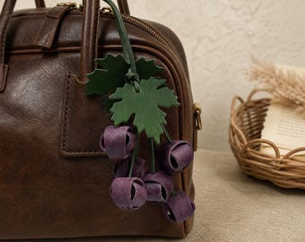 Puede incluir: Un bolso de mano de cuero marrón oscuro con un adorno decorativo en forma de racimo de uvas. El adorno consta de varias esferas de cuero morado que representan uvas, unidas a un tallo verde con dos hojas de parra de cuero verde.