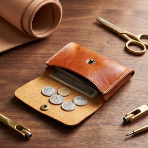 May include: A tan leather wallet with a snap button closure, containing US paper currency and five silver-colored coins resting on the open flap. The wallet features visible white stitching along the edges and a smooth, distressed leather texture.