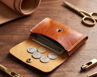 Puede incluir: Una cartera de cuero color canela con cierre de botón a presión, que contiene billetes y cinco monedas de color plateado sobre la solapa abierta. La cartera presenta costuras blancas visibles en los bordes y una textura de cuero suave.