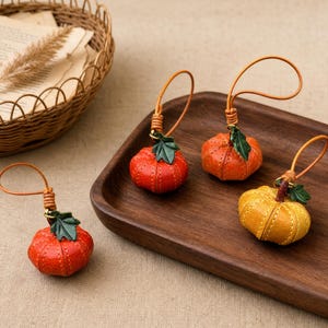 Puede incluir: Tres pequeños dijes de cuero con forma de calabaza con detalles cosidos, cada uno con un acento de hoja verde y un lazo de cuero fino para colgar. Dos dijes son de color naranja vibrante y uno es de un color amarillo dorado más claro.
