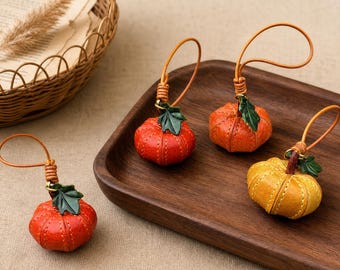 Puede incluir: Tres pequeños dijes de cuero con forma de calabaza con detalles cosidos, cada uno con un acento de hoja verde y un lazo de cuero fino para colgar. Dos dijes son de color naranja vibrante y uno es de un color amarillo dorado más claro.