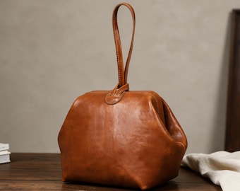 Puede incluir: Un bolso de mano de cuero color canela con una abertura de marco estructurado y una sola asa vertical superior. El bolso presenta un acabado liso y pulido con costuras visibles a lo largo de los bordes y un cierre en la parte superior central.