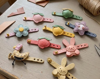Puede incluir: Una colección de tiradores de cuero para cajones con diversas formas, incluyendo una flor azul claro, un melocotón rosa, una fruta verde, un cítrico amarillo, una estrella morada, una fruta roja, un conejo rosa, un conejo amarillo y un pájaro amarillo con detalles negros.