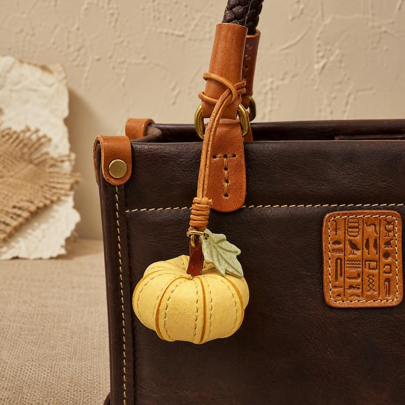 Puede incluir: Un bolso de cuero marr&oacute;n oscuro con un parche cuadrado con jerogl&iacute;ficos egipcios en relieve y un dije colgante con forma de calabaza amarilla con una peque&ntilde;a hoja verde. El bolso tiene detalles en cuero marr&oacute;n claro y herrajes de lat&oacute;n.