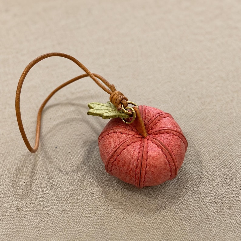 Puede incluir: Un peque&ntilde;o dije con forma de calabaza de color rosa rojizo y textura, elaborado con costuras visibles. Presenta un detalle de hoja verde claro unido a un anillo de lat&oacute;n y un lazo de cord&oacute;n de cuero color canela para colgar, creando un adorno en miniatura.