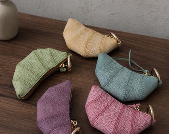 Puede incluir: Cinco monederos en forma de media luna en colores beige, verde salvia, morado, azul polvoriento y rosa polvoriento. Cada monedero presenta una superficie texturizada, una cremallera dorada y una anilla metálica dorada en un extremo.