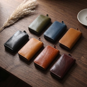 Könnte beinhalten: Eine Sammlung von sieben rechteckigen Reißverschlusstaschen in verschiedenen Farben, darunter Olivgrün, Marineblau, Schwarz, Hellbraun, Mittelbraun und Dunkelbraun. Jede Tasche hat eine glatte Oberfläche, abgerundete Ecken und eine kleine Zuglasche.