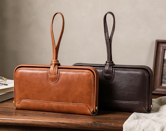 Puede incluir: Dos carteras rectangulares con correa de muñeca, una de color tostado y otra marrón oscuro, cada una con un acabado de piel lisa, una estructura rígida y una correa vertical sujeta en la parte superior central. Diseño limpio y sencillo.