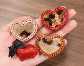 Puede incluir: Cuatro accesorios de cuero en forma de corazón con bordes cosidos. Tres corazones presentan incrustaciones de piel con estampado animal en tonos marrones y negros. Un corazón es de cuero rojo liso con un pequeño lazo de cuero negro en el lateral.