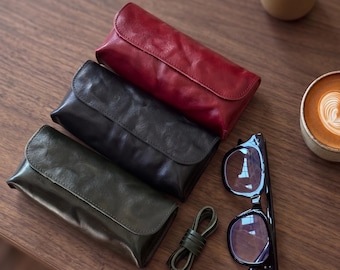 Puede incluir: Tres estuches rectangulares de cuero con solapas plegables en rojo, negro y verde oscuro. Junto a los estuches hay un pequeño cordón de cuero enrollado y unas gafas de montura negra con lentes transparentes, situadas cerca de un café.