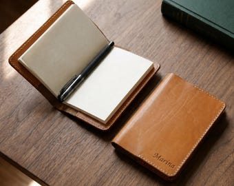 Puede incluir: Dos fundas de cuero color canela con costuras visibles. Una funda está abierta, mostrando páginas blancas en blanco y un bolígrafo negro en su interior. La otra funda está cerrada y presenta el nombre Marina grabado en la esquina inferior.