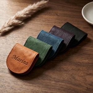 Könnte beinhalten: Eine Reihe von fünf kleinen, U-förmigen Münztaschen aus Leder in den Farben Hellbraun, Grün, Blau, Violett und Schwarz. Die hellbraune Tasche im Vordergrund ist mit dem Namen Marina in Schreibschrift geprägt. Jede Tasche hat sichtbare Nähte.