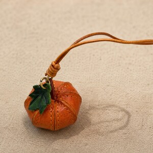 Puede incluir: Un peque&ntilde;o dije de calabaza de cuero naranja con costuras verticales y una hoja de cuero verde sujeta a una argolla de lat&oacute;n. El dije cuelga de un cord&oacute;n de cuero fino de color marr&oacute;n claro con un detalle de nudo.
