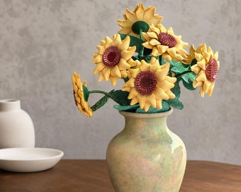 Puede incluir: Un ramo de girasoles artificiales con pétalos amarillos y centros de color rojo oscuro, con tallos y hojas verdes, dispuesto en un jarrón de cerámica redondeado de tonos pastel moteados, con acabado liso y cuello estrecho.