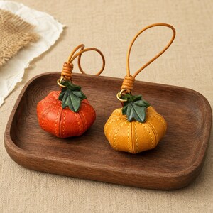 Puede incluir: Dos colgantes de cuero con forma de calabaza y detalles de costura, cada uno con una hoja de cuero verde y un fino lazo de cuero para colgar. Una calabaza es de color naranja vibrante y la otra es de color amarillo dorado, ambas con textura.