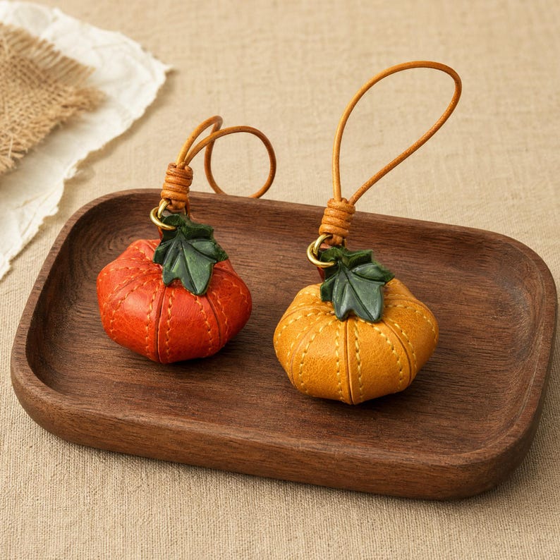 Puede incluir: Dos colgantes de cuero con forma de calabaza y detalles de costura, cada uno con una hoja de cuero verde y un fino lazo de cuero para colgar. Una calabaza es de color naranja vibrante y la otra es de color amarillo dorado, ambas con textura.
