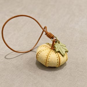 Puede incluir: Un peque&ntilde;o dije con forma de calabaza de color amarillo p&aacute;lido con costuras visibles, que presenta un acento de hoja verde texturizada unido a un cord&oacute;n de cuero marr&oacute;n fino. La calabaza tiene una forma redondeada y segmentada con un anillo de metal.