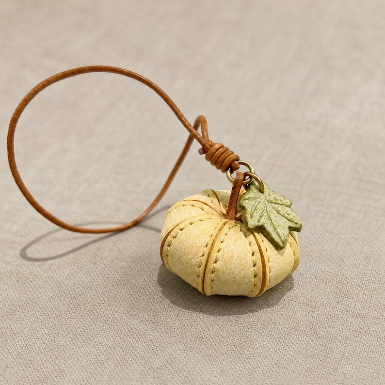 Puede incluir: Un peque&ntilde;o dije con forma de calabaza de color amarillo p&aacute;lido con costuras visibles, que presenta un acento de hoja verde texturizada unido a un cord&oacute;n de cuero marr&oacute;n fino. La calabaza tiene una forma redondeada y segmentada con un anillo de metal.