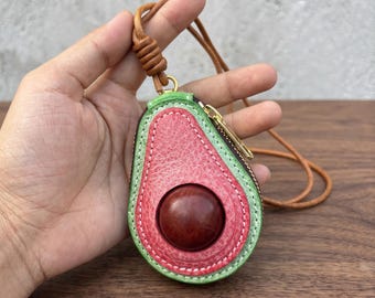 Puede incluir: Un estuche de cuero con forma de mitad de aguacate, con un centro rosa texturizado, un acento circular liso de color marrón oscuro y un borde verde con costuras blancas. Incluye una cremallera dorada y un cordón largo y fino de cuero color canela.