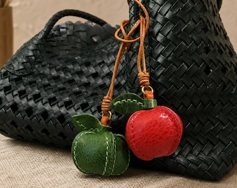 Puede incluir: Dos bolsos negros trenzados con un par de colgantes de fruta. Un colgante es una manzana roja texturizada y el otro una manzana verde texturizada, ambos con hojas verdes y sujetos a los bolsos mediante finos cordones de cuero color canela.