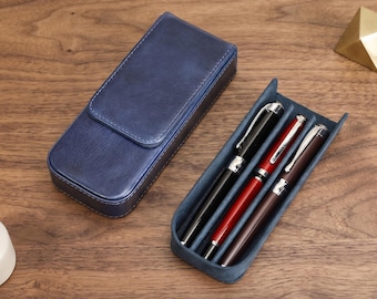 Puede incluir: Un estuche rectangular para bolígrafos de color azul marino con cierre de solapa, acompañado de una bandeja abierta que contiene tres bolígrafos: uno negro, uno rojo y uno marrón oscuro, cada uno con clip y detalles plateados.