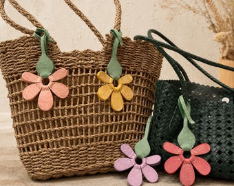 Puede incluir: Un bolso tote de paja trenzada en color tostado natural y un bolso de cuero trenzado verde oscuro, ambos con asas trenzadas. Los bolsos llevan colgantes de cuero en forma de flor en tonos pastel rosa, amarillo, lavanda y coral con tallos verdes.