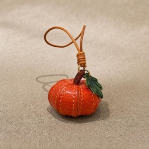 Puede incluir: Un peque&ntilde;o dije de calabaza de cuero naranja texturizado con costuras visibles de color claro, un tallo marr&oacute;n oscuro y una hoja verde. La calabaza est&aacute; unida a un lazo de cord&oacute;n de cuero color canela con un nudo de cuero enrollado.