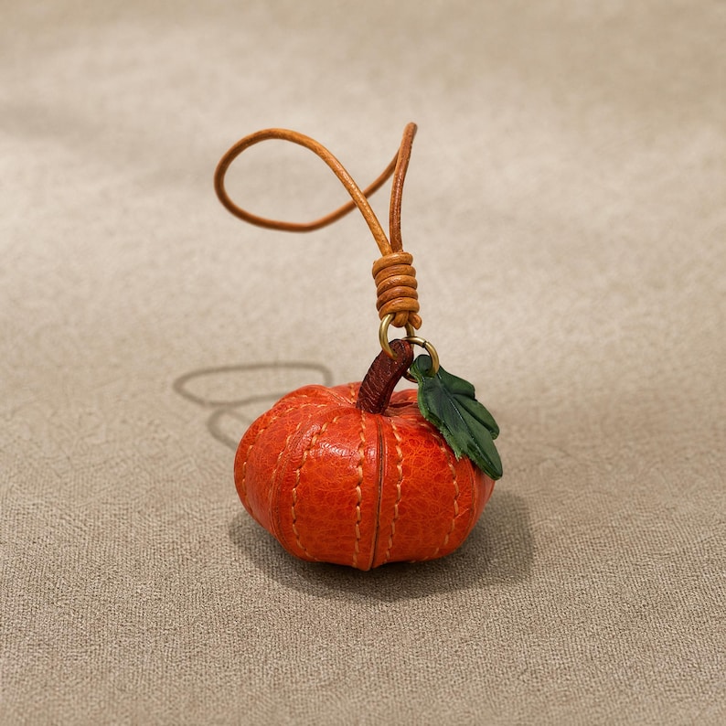 Puede incluir: Un peque&ntilde;o dije de calabaza de cuero naranja texturizado con costuras visibles de color claro, un tallo marr&oacute;n oscuro y una hoja verde. La calabaza est&aacute; unida a un lazo de cord&oacute;n de cuero color canela con un nudo de cuero enrollado.