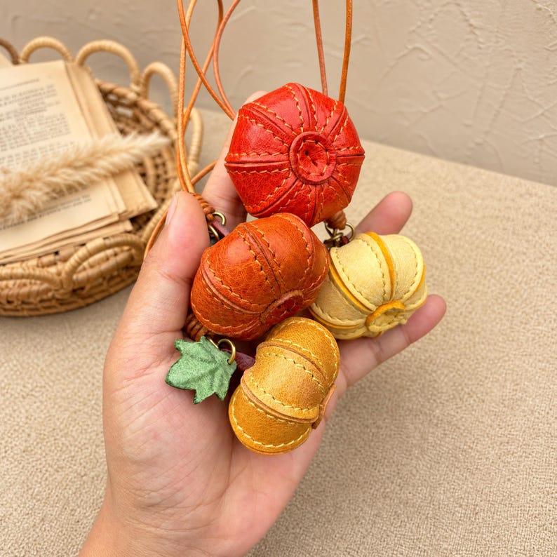 Puede incluir: Un adorno colgante decorativo que presenta cuatro peque&ntilde;as calabazas de cuero cosido en tonos rojo, naranja, amarillo y dorado, acentuadas con un dije de hoja de cuero verde. Las calabazas est&aacute;n suspendidas de finos cordones de cuero marr&oacute;n.