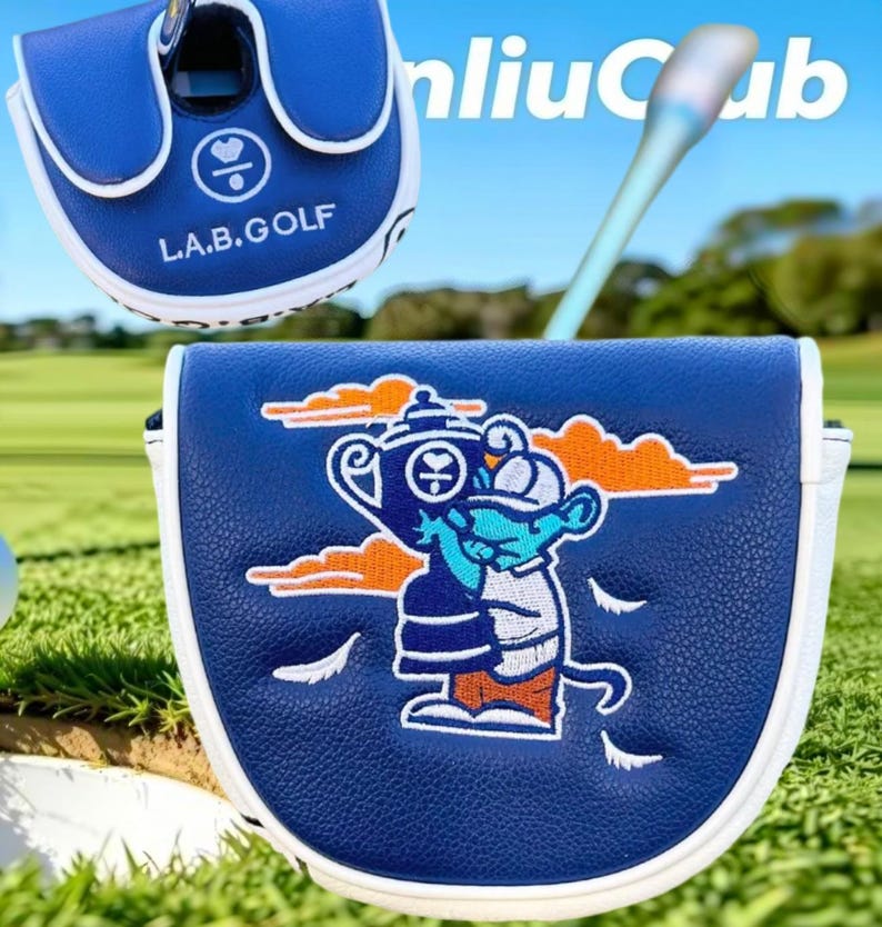 May include: A blue and white golf putter headcover featuring the text L.A.B.GOLF and an embroidered graphic of a cartoon character holding a trophy. The design includes orange clouds and white feathers on a textured blue surface with white trim.