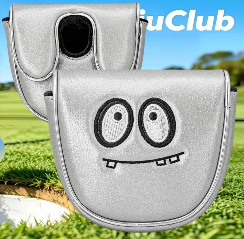 May include: A silver mallet putter headcover featuring an embroidered cartoon face with two large oval eyes and a crooked mouth with visible teeth. The cover has a smooth metallic finish and a black interior lining. The text "iuClub" is visible.