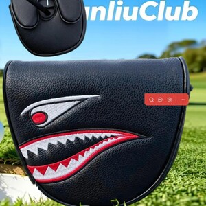 May include: A black mallet putter headcover featuring a textured leather-like finish and an embroidered shark mouth graphic with white teeth and red accents. The design includes a stylized eye with a red pupil. The text "nliuClub" is visible.