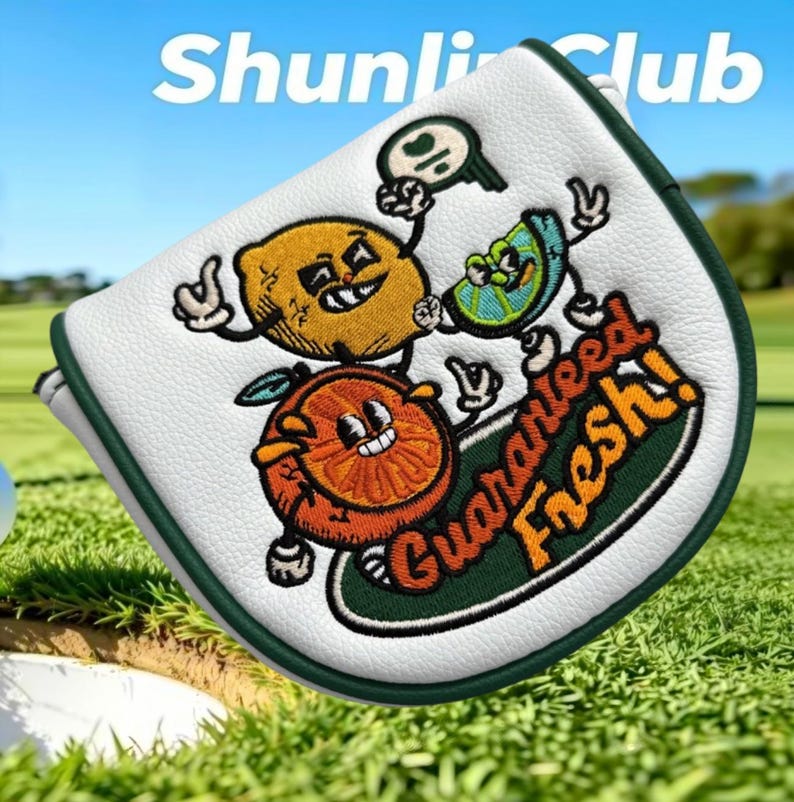 Puede incluir: Una funda para putter de golf blanca con borde verde oscuro que presenta un gr&aacute;fico bordado de un lim&oacute;n amarillo, una lima verde y una rodaja de naranja con caras y extremidades de dibujos animados. El texto "Guaranteed fresh!" est&aacute; bordado en naranja.